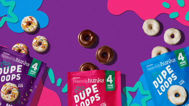 Three bags of Heavenly Hunks Dupe Loops mini donuts are arranged on a bright purple background with colorful abstract shapes and scattered real mini donuts. The bags are purple, pink, and blue, and the donuts include chocolate glaze and fruit topping flavors.